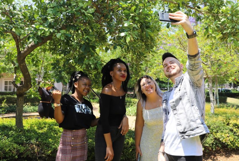 Four students taking a selfie together under a shaded tree on AAA School of Advertising campus